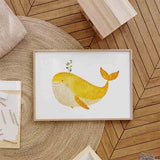 Cuadro horizontal con ballena amarilla de gran tamaño, vientre crema, brote verde y fondo blanco, en composición suave de estilo infantil. Disponible en kudeko.com