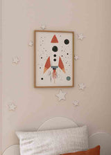 Quadro emoldurado com foguete espacial infantil de formas simples, fundo claro, pequenos pontos pretos, planetas discretos, tons vermelho tijolo, cinzento azulado e bege, estilo infantil nórdico. Disponível em kudeko.com