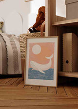 Quadro emoldurado com baleia branca sobre mar azul, céu coral, sol claro e formas suaves num estilo infantil nórdico sereno. Disponível em kudeko.com