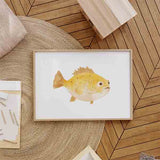 Quadro horizontal com peixe infantil em amarelo dourado e creme, pequenos detalhes castanhos, fundo branco e estilo de ilustração suave e alegre para quarto de bebé ou infantil. Disponível em kudeko.com