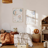 Quadro infantil com girafa sobre lua azul, leão astronauta, estrelas em tons pastel e planeta celeste sobre fundo branco, em estilo infantil suave com azul, rosa, bege e cinzento. Disponível em kudeko.com