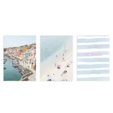 Conjunto de três quadros emoldurados com porto costeiro, casas em tons pastel, praia de areia clara com banhistas, tipografia rosa sobre riscas em azul suave e estilo fotográfico luminoso. Disponível em kudeko.com