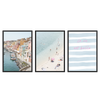 Conjunto de três quadros emoldurados com porto costeiro, casas em tons pastel, praia de areia clara com banhistas, tipografia rosa sobre riscas em azul suave e estilo fotográfico luminoso. Disponível em kudeko.com