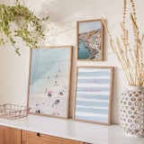Quadro com vista aérea de praia com areia branca, água turquesa clara, banhistas dispersos, guarda-sóis em tons suaves e estilo fotográfico luminoso. Disponível em kudeko.com