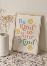 Tableau avec la phrase Be Kind to Your Mind, grandes lettres multicolores, visages souriants jaunes, fond clair et style typographique pastel moderne. Disponible sur kudeko.com