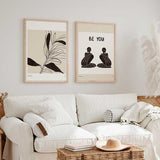 Quadro emoldurado com frase Be You, duas figuras femininas sentadas a preto sobre fundo bege claro, composição simétrica em estilo de ilustração simples. Disponível em kudeko.com