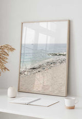 Cuadro de playa luminosa con arena beige, orilla de piedras claras, mar azul suave y velero lejano, de estilo fotográfico. Disponible en kudeko.com