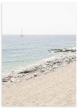 Cuadro de playa luminosa con arena beige, orilla de piedras claras, mar azul suave y velero lejano, de estilo fotográfico. Disponible en kudeko.com