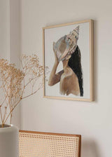 Quadro emoldurado com figura feminina de perfil a segurar uma grande concha marinha sobre o rosto, em tons de bege, areia, marfim, cinzento suave e preto, com estilo de ilustração costeiro. Disponível em kudeko.com