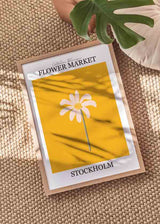 Gerahmtes Wandbild mit zentrierter weißer Blüte, senfgelbem Hintergrund, schwarzer Schrift Flower Market Stockholm und klarer floraler Gestaltung im grafischen Stil. Erhältlich auf kudeko.com