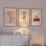 Quadro com menina sentada sobre uma arca castanha, casaco amarelo, vestido floral, fundo bege e estilo ilustrado suave para quarto infantil. Disponível em kudeko.com
