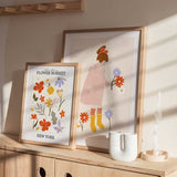 Cuadro de ilustración infantil con niña de vestido rosa, lazo rojo, botas amarillas y flores lila, naranja y blancas sobre fondo beige suave. Disponible en kudeko.com