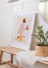 Cuadro de ilustración infantil con niña de vestido rosa, lazo rojo, botas amarillas y flores lila, naranja y blancas sobre fondo beige suave. Disponible en kudeko.com