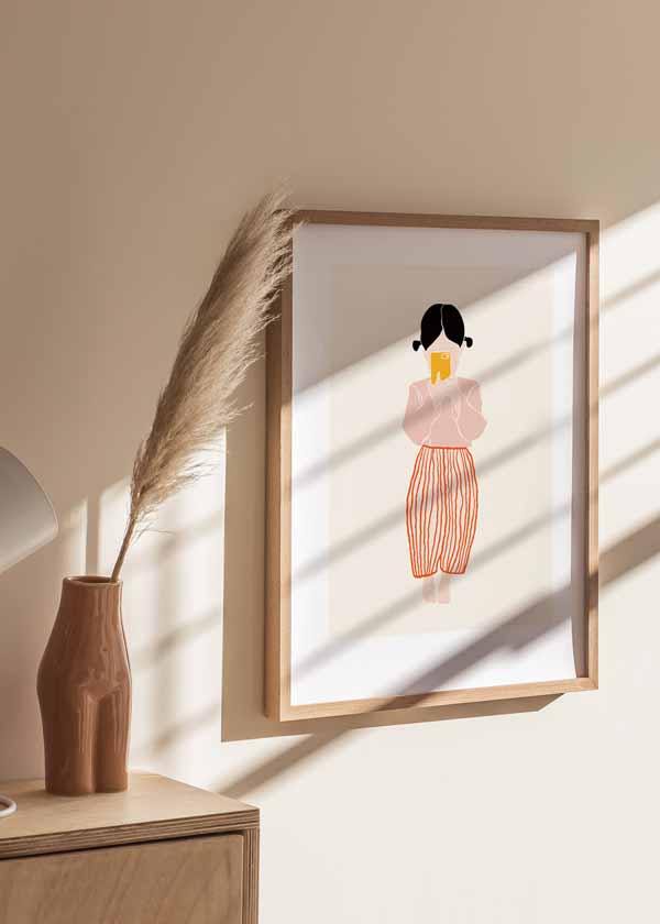 Tableau encadré avec fillette aux cheveux noirs, blouse rose, pantalon à rayures orange, mobile jaune, fond crème et style enfantin aux formes douces. Disponible sur kudeko.com