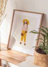 Gerahmtes Wandbild mit Mädchen, gelbem Hut, Sonnenbrille, rosa Blumenbluse, brauner Tasche, hellem beigem Hintergrund und schlichtem kindlichem Stil. Erhältlich auf kudeko.com