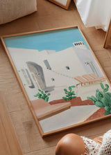 Quadro de arquitetura mediterrânica com casas brancas, céu azul, espreguiçadeiras bege, cactos verdes em primeiro plano e estilo natural de paisagem serena. Disponível em kudeko.com