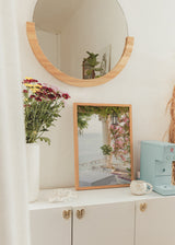 Wandbild einer mediterranen Terrasse am Meer mit rosa Bougainvillea, weißen Säulen, schwarzer Laterne, grüner Vegetation und sanftem Licht im fotografischen Stil. Erhältlich auf kudeko.com