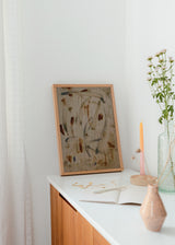 Quadro emoldurado com formas abstratas orgânicas sobre fundo bege com textura visual de madeira, em tons de castanho ferrugem, areia e cinzento azulado, de estilo abstrato contemporâneo. Disponível em kudeko.com