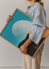 Gerahmtes Wandbild mit großem weißem Mond, türkisfarbenem Himmel, orangefarbener Düne, einsamer Silhouette und dunkler Landschaft im fotografischen surrealen Stil. Erhältlich auf kudeko.com