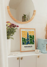 Gerahmtes Wandbild mit Burj Khalifa in Dunkelblau, grünen Blättern, gelben organischen Formen, hellem Hintergrund und urbanem Illustrationsstil mit klarer Typografie. Erhältlich auf kudeko.com
