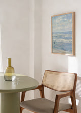 Quadro emoldurado de praia com ondas suaves, céu nublado, figura solitária junto à beira-mar, em azul-claro, branco e cinzento, de estilo vintage. Disponível em kudeko.com