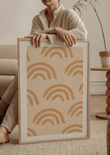 Quadro emoldurado com arco-íris repetidos em bege, areia e terracota suave sobre fundo creme, num estilo infantil moderno e equilibrado para quarto de bebé ou canto de brincadeira. Disponível em kudeko.com