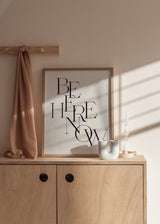 Gerahmtes Wandbild mit dem Schriftzug Be Here Now in schwarzer Serifentypografie auf hellbeigem Hintergrund, klare Komposition im zeitgemäßen Textstil mit ausgewogener Wirkung. Erhältlich auf kudeko.com