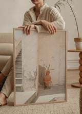 Quadro de um recanto mediterrânico com escada de pedra, parede branca envelhecida, porta clara, peça de terracota, planta verde e estilo fotográfico em tons de creme, areia e terracota. Disponível em kudeko.com