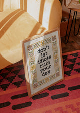 Cuadro decorativo de estilo minimalista y orgánico colocado en salón, frase bordada con humor y marco floral, modelo Don't Let Idiots Quote