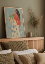 Tableau encadré avec femme de profil tenant une plante en pot, fond bleu gris doux, robe crème à fleurs bleues, blouse terracotta, style illustration décorative. Disponible sur kudeko.com