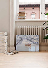 Quadro horizontal com três veados sobre neve branca, sob grandes ramos cobertos de geada, em tons de branco, cinzento suave e castanho claro, de estilo fotográfico invernal. Disponível em kudeko.com