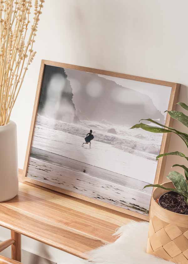 Horizontales Wandbild mit einem Surfer auf dem Weg ins Meer, zwischen weißen Wellen, nassem Sand, grauen Klippen, sanften Beigetönen und weichem Licht im fotografischen Stil. Erhältlich auf kudeko.com