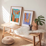 Cuadro de dos manos entrelazadas con flores rosadas, margaritas blancas, campanillas azules y fondo azul suave, en estilo ilustración. Disponible en kudeko.com