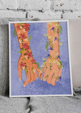 Cuadro de dos manos entrelazadas con flores rosadas, margaritas blancas, campanillas azules y fondo azul suave, en estilo ilustración. Disponible en kudeko.com