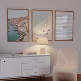 Conjunto de três quadros emoldurados com porto costeiro, casas em tons pastel, praia de areia clara com banhistas, tipografia rosa sobre riscas em azul suave e estilo fotográfico luminoso. Disponível em kudeko.com