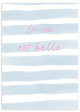 Quadro emoldurado com frase La vie est belle, riscas horizontais em azul céu e branco, tipografia rosa suave e estilo tipográfico contemporâneo para quarto ou zona de toucador. Disponível em kudeko.com