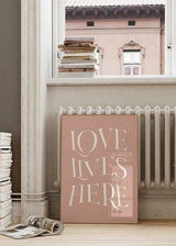 Cuadro con frase LOVE LIVES HERE en letras blancas grandes sobre fondo rosa empolvado, composición tipográfica elegante de estilo con frases. Disponible en kudeko.com