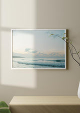 Quadro horizontal de mar calmo ao amanhecer com ondas suaves, céu claro, nuvens difusas, silhueta ao longe, tons azul-claro, bege e branco, estilo fotográfico. Disponível em kudeko.com