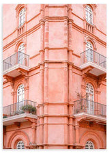 Cuadro de fachada clásica rosa pastel con balcones de hierro, ventanas arqueadas y detalles ornamentales, de estilo fotográfico. Disponible en kudeko.com