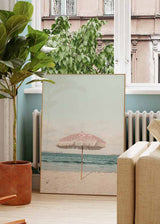 Cuadro de sombrilla rosa con flecos sobre playa de arena clara, mar azul sereno y cielo pastel, en estilo fotográfico. Disponible en kudeko.com