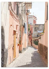 Quadro emoldurado de uma rua estreita mediterrânica com fachadas em rosa, bege e terracota, chão empedrado, varandas, roupa estendida, luz suave e estilo fotográfico. Disponível em kudeko.com