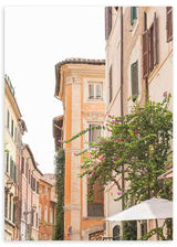 Gerahmtes Wandbild einer römischen Straße mit Fassaden in Creme, Pfirsich und Sand, dunklen Fensterläden, Balkonen, grüner Vegetation, fuchsiafarbenen Blüten und mediterranem Stadtstil. Erhältlich auf kudeko.com