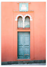 Fotografisches Wandbild mit verwitterter blauer Tür auf korallfarbener Fassade, Fenster mit doppelten Bögen, türkisen und weißen Details, symmetrischer Komposition und mediterranem Architekturstil. Erhältlich auf kudeko.com