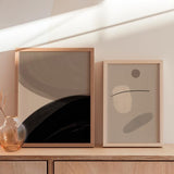 Quadro abstrato emoldurado com formas orgânicas bege, cinzento taupe, castanho suave e linha preta curva, num estilo contemporâneo equilibrado. Disponível em kudeko.com