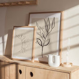 Wandbild mit botanischem Zweig, länglichen schwarzen Blättern auf hellbeigem Hintergrund, pflanzliche Komposition im minimalistischen Stil mit neutralen Farbtönen. Erhältlich auf kudeko.com