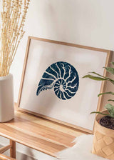 Quadro horizontal com concha fóssil em espiral tipo amonite, em azul-marinho sobre fundo bege envelhecido, composição centrada e estilo vintage de inspiração costeira. Disponível em kudeko.com