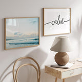 Quadro horizontal de mar calmo ao amanhecer com ondas suaves, céu claro, nuvens difusas, silhueta ao longe, tons azul-claro, bege e branco, estilo fotográfico. Disponível em kudeko.com