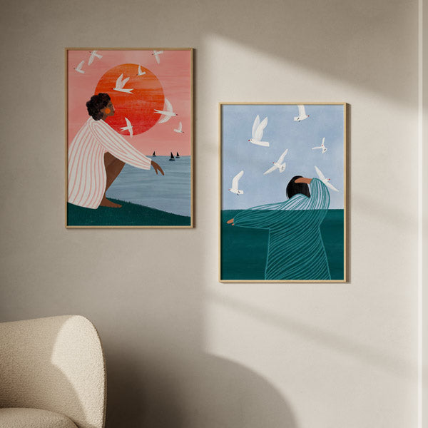 Set aus zwei gerahmten Wandbildern mit Frauen am Meer, weißen Vögeln im Flug, roter Sonne, rosa und blauem Himmel, im Illustrationsstil mit türkisen und schwarzen Farbtönen. Erhältlich auf kudeko.com