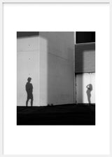 Cuadro de sombras de dos personas sobre pared urbana de hormigón y puerta metálica, en tonos blanco, negro y gris, de estilo fotográfico. Disponible en kudeko.com