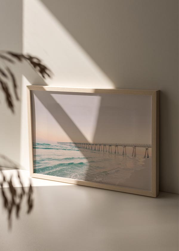 Cuadro horizontal de un muelle sobre el mar con olas suaves, arena clara, cielo rosado y tonos turquesa, de estilo fotográfico. Disponible en kudeko.com